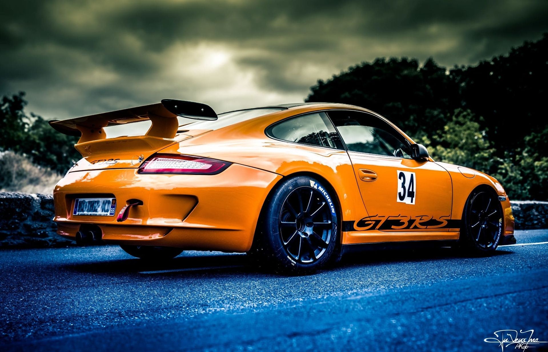 Seitliche Ansicht eines orangefarbenen Sportwagens mit Heckspoiler auf einer nassen Straße unter bewölktem Himmel