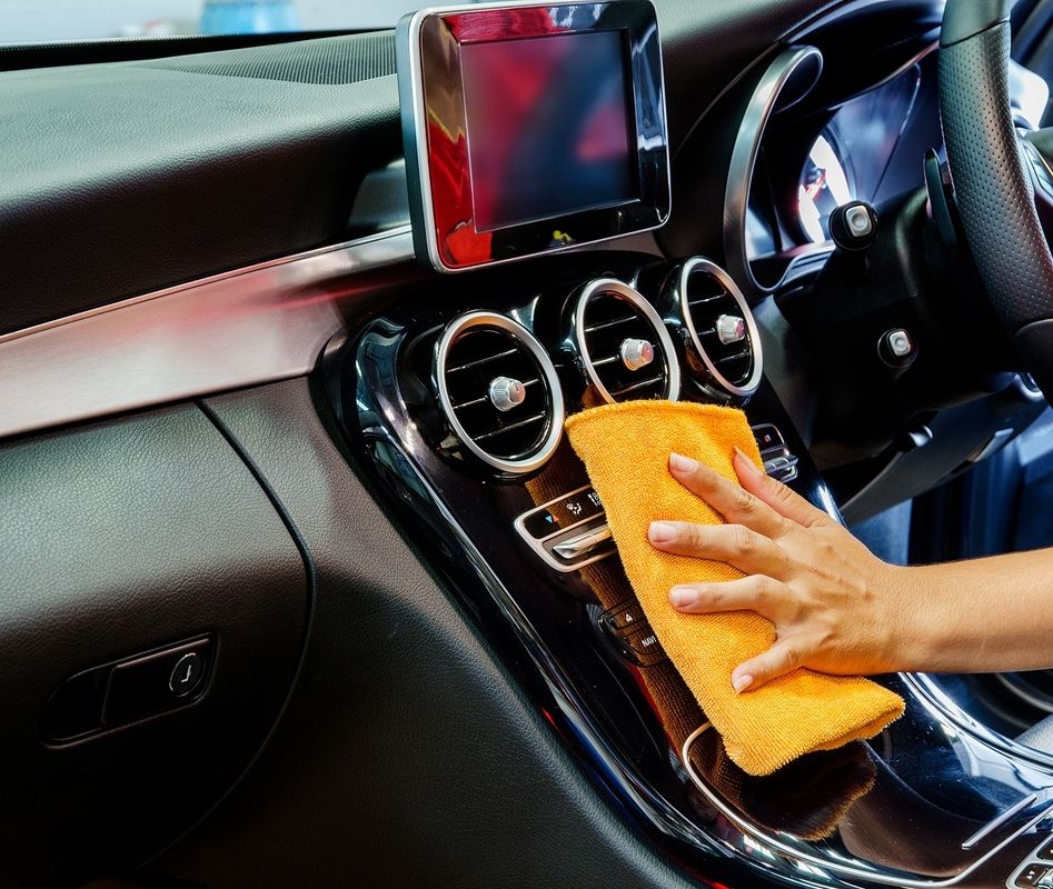 Hand wischt mit gelbem Tuch die Mittelkonsole eines Autos mit drei runden Lüftungsdüsen und einem Display ab