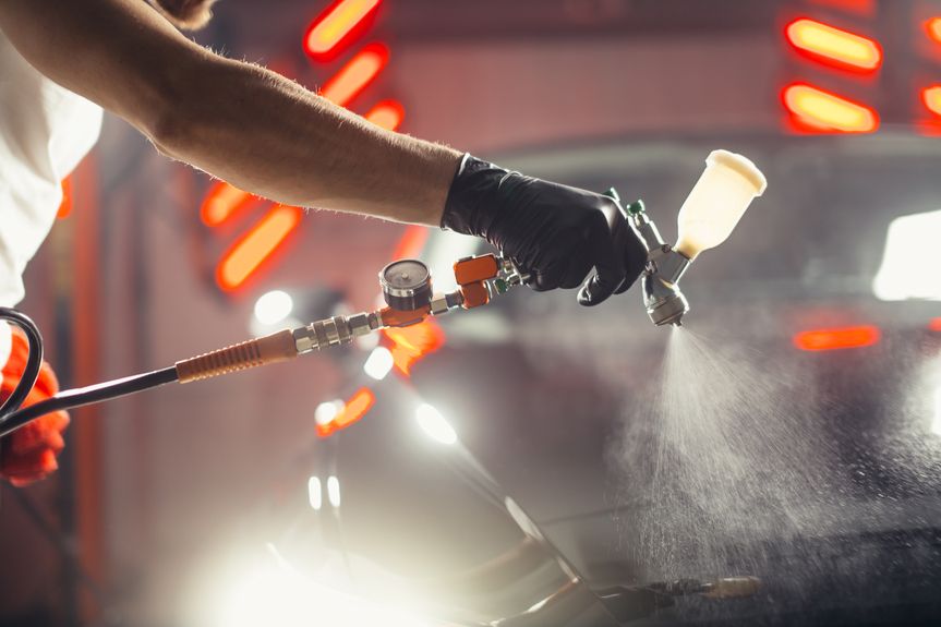 Hand mit schwarzem Handschuh hält eine Spritzpistole, die feinen Nebel auf eine Oberfläche sprüht